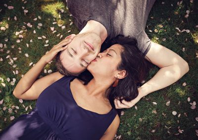 Family photographer Ania Chandra in REading area. Married couple are lying on the grass in Harris gardens in Reading during spring time and wife is kissing her husband in his cheek - they are lying face to face - they both have eyes closed :)