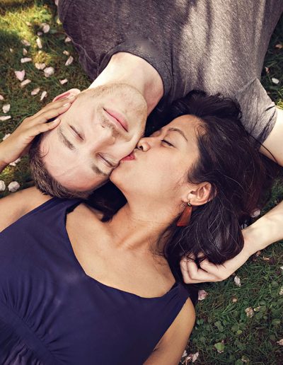 Family photographer Ania Chandra in REading area. Married couple are lying on the grass in Harris gardens in Reading during spring time and wife is kissing her husband in his cheek - they are lying face to face - they both have eyes closed :)