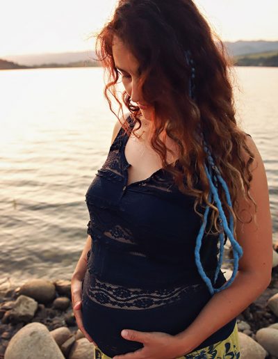 Ania Chandra maternity photographer in Berkshire and beyond. Photo of pregnant mother during sunset on the lake. She has long and curly black hair and she is looking down on her belly while holding her tommy with both hands.