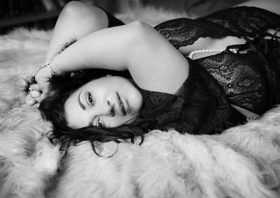 Curvie women with black hair and beautiful big eyes is lying on the bed with hands up and facing camera - smiling ever so gently - Boudoir photographer Reading -Ania chandra photographer