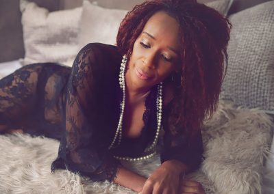 black beautiful women is lying down in lace gown with perl neckless on her neck and she is looking down - Boudoir image done by Ania Chandra photographer in Reading