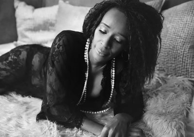 Beautiful black women is lying on her front looking down - dressed in black gown and purl neckless on her neck - image done by Boudoir Ania Chandra Photography in Rearing